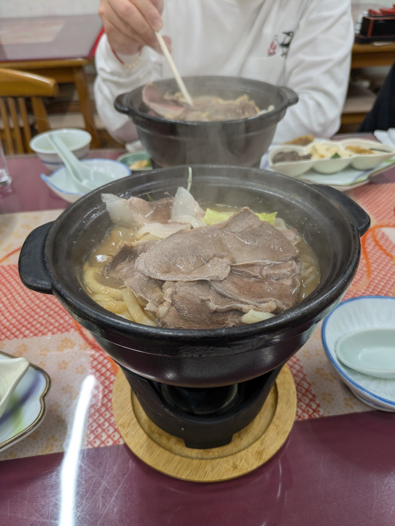 一人用コンロで湯気を立てる猪鍋。肉の下にうどん、奥でも箸で鍋をつつく人