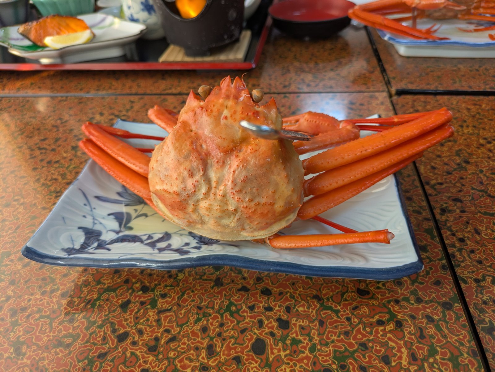 漆塗りの座卓の染付皿に盛られた丸ごと一杯の茹で蟹、奥に焼き鮭や固形燃料の鍋