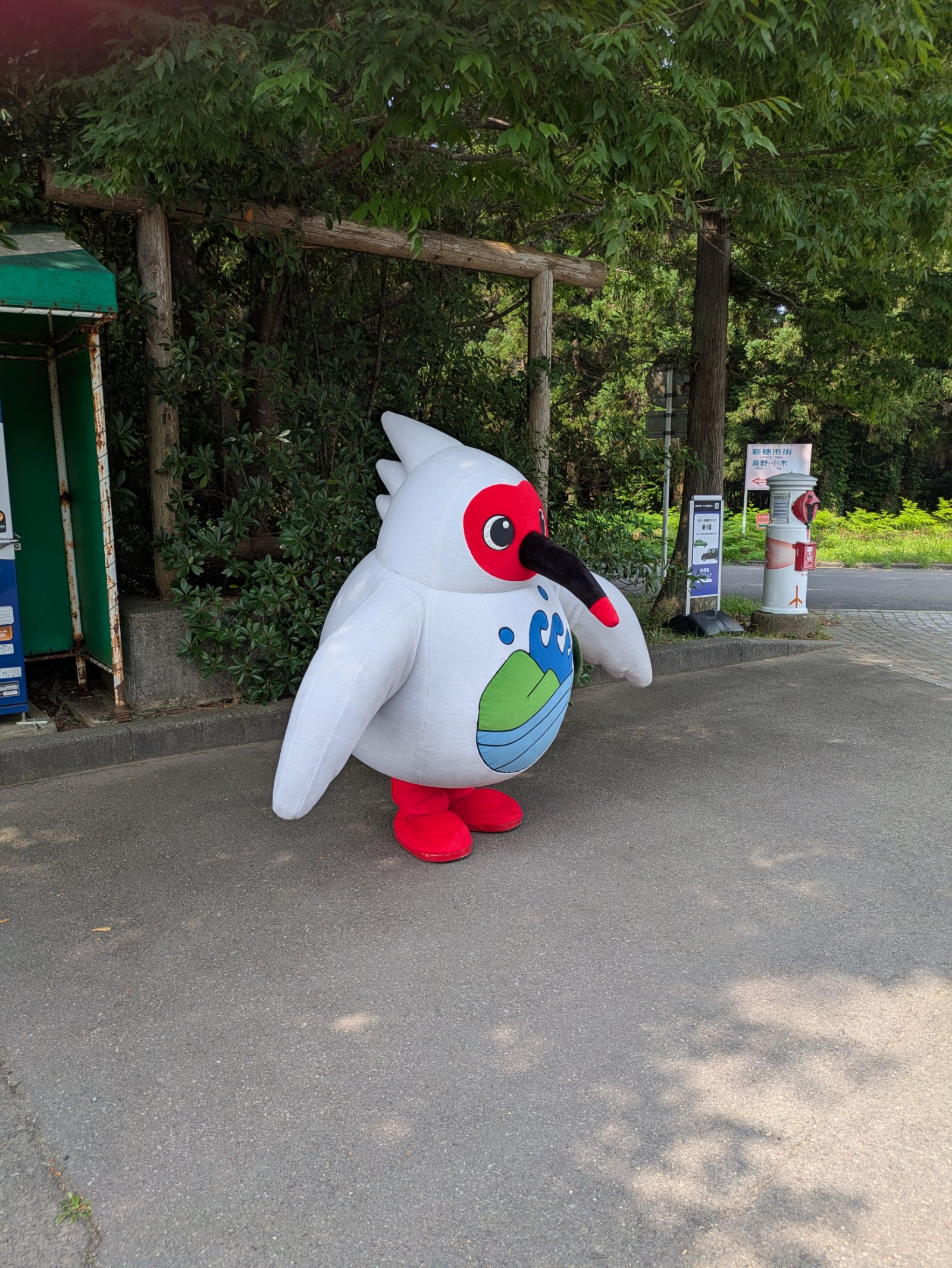 夏の木陰の駐車場に立つトキの着ぐるみキャラクター、奥にトキの郵便ポスト