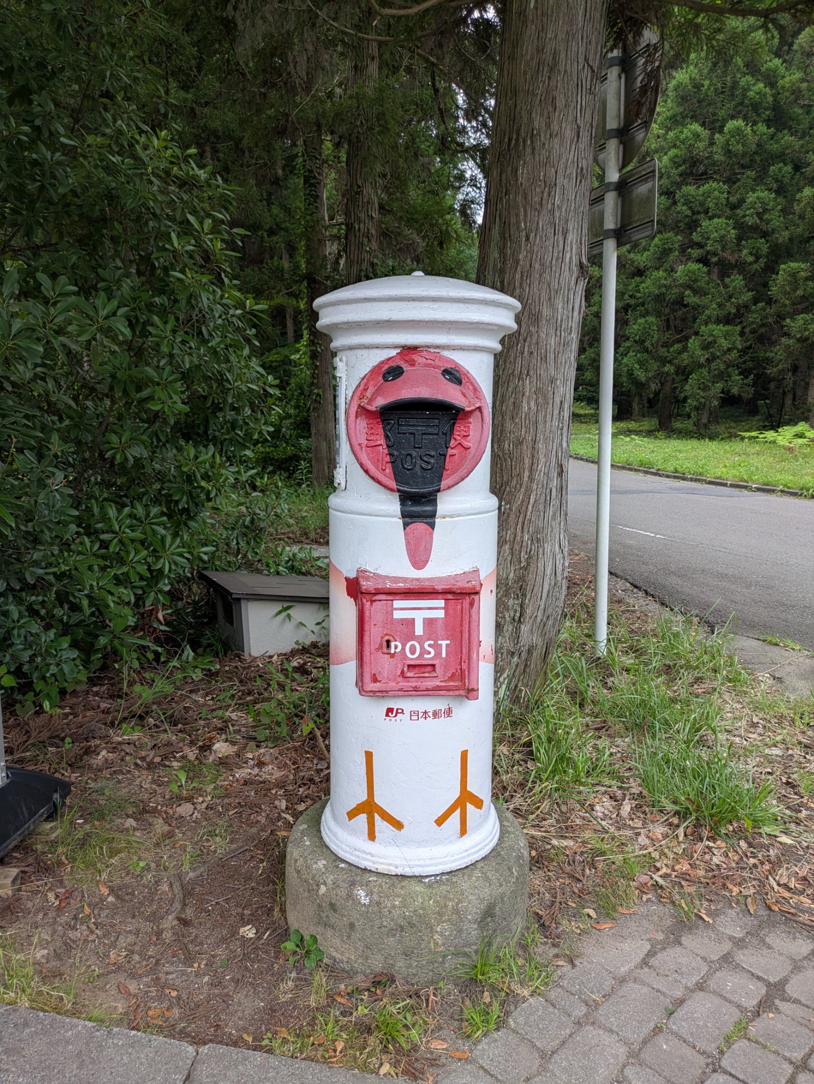 トキの顔と足が描かれた白い円筒形の郵便ポスト、佐渡のトキの森公園の木立の脇