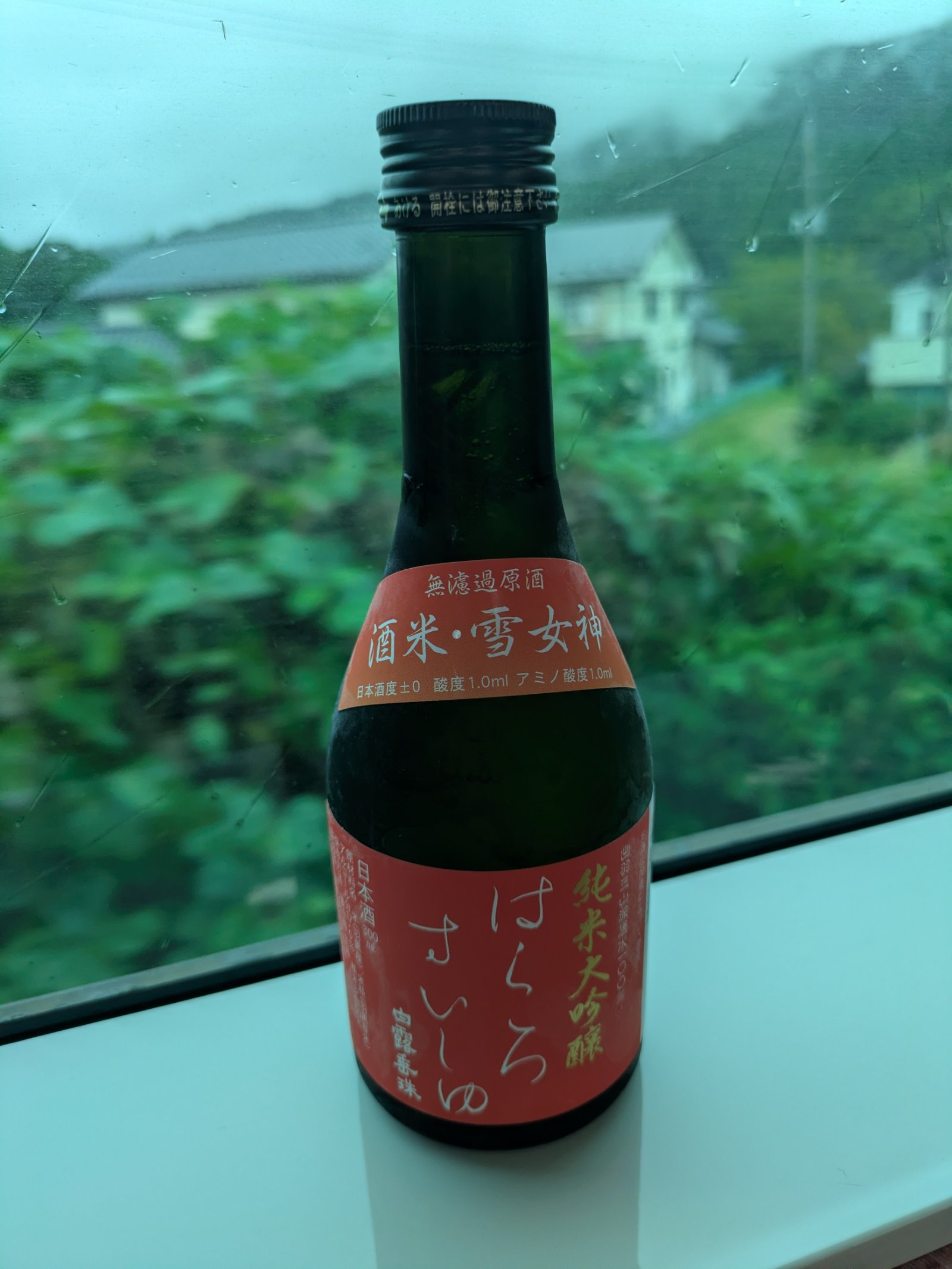 雨の車窓に置かれた純米大吟醸「はくろすいしゅ」の小瓶、外には緑の木々と民家