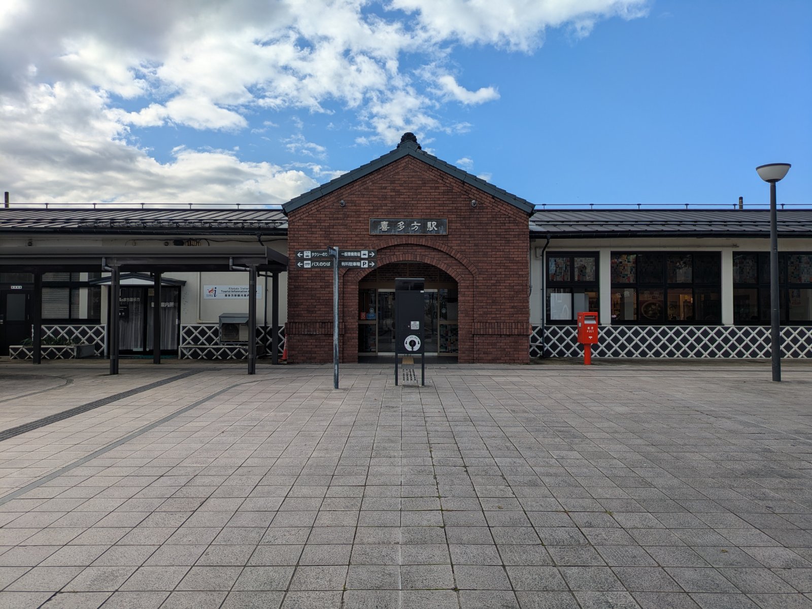 赤レンガの切妻造りの入口に「喜多方駅」の看板が掲げられた駅舎と駅前広場、晴れ間ののぞく空