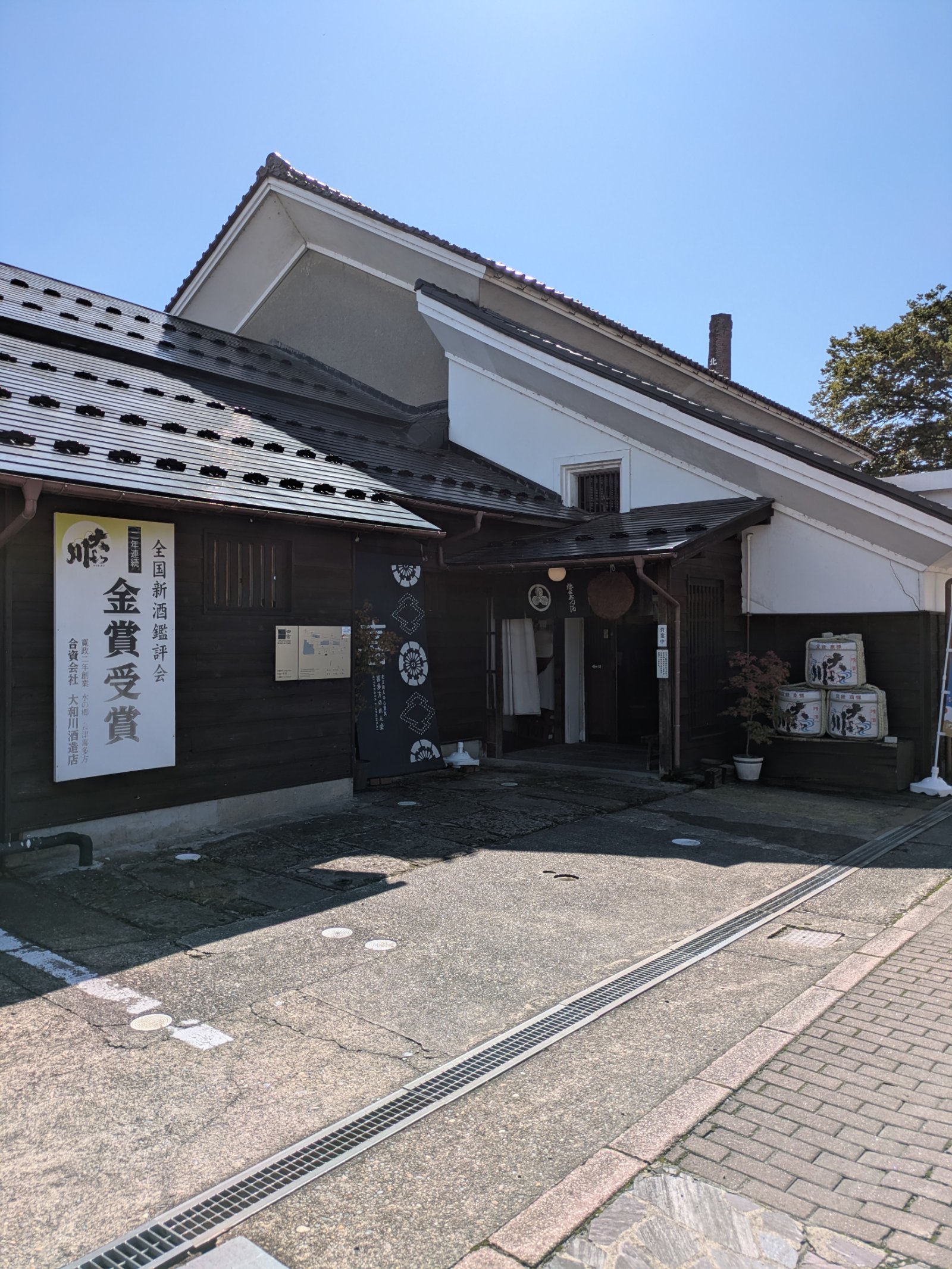 白壁の蔵と黒い板壁が並ぶ大和川酒造店の入口。全国新酒鑑評会金賞受賞の看板と酒樽が置かれ、青空が広がる