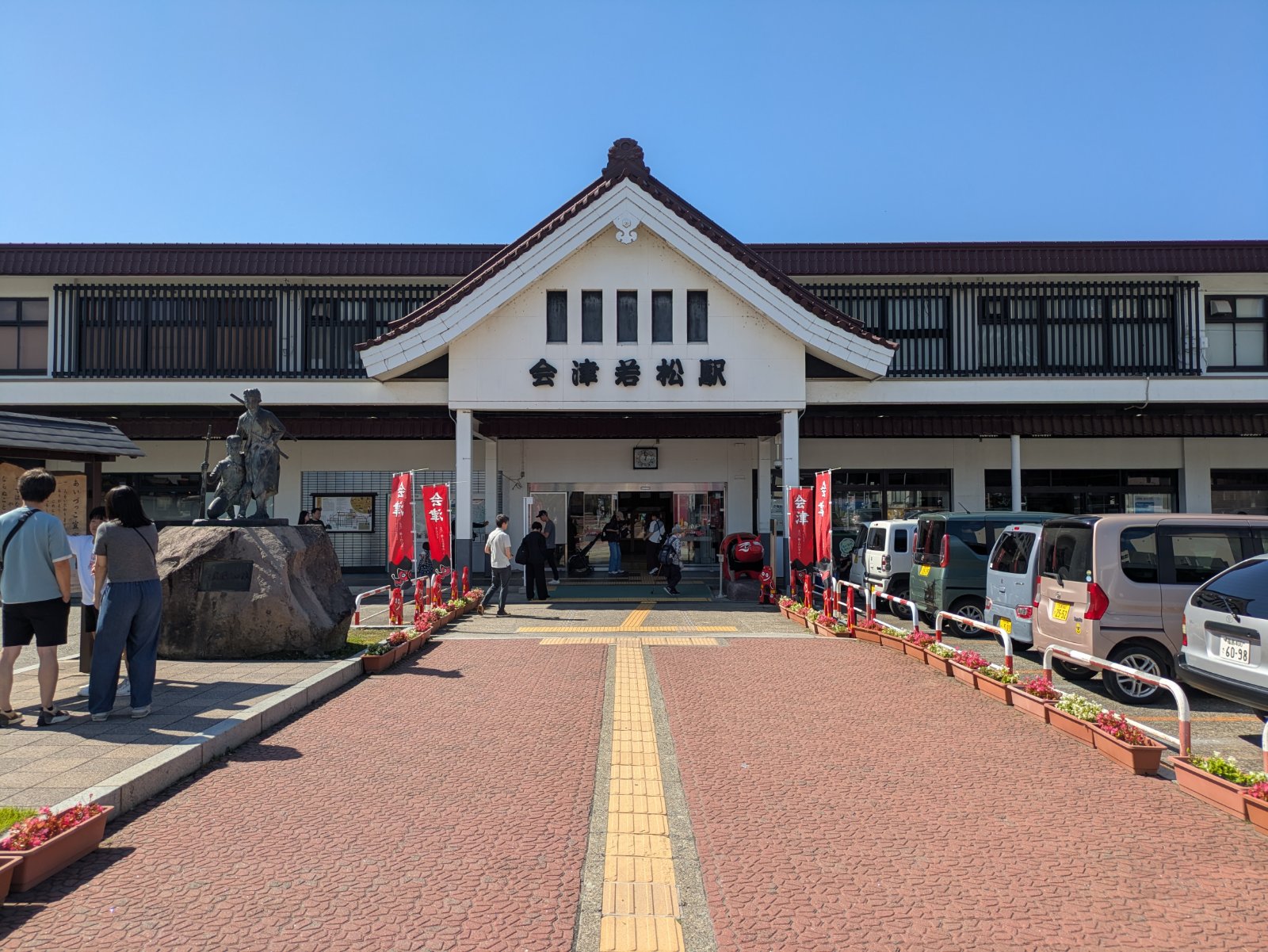 晴天の下の会津若松駅の駅舎。赤いのぼりが並ぶ入口と駅前の銅像