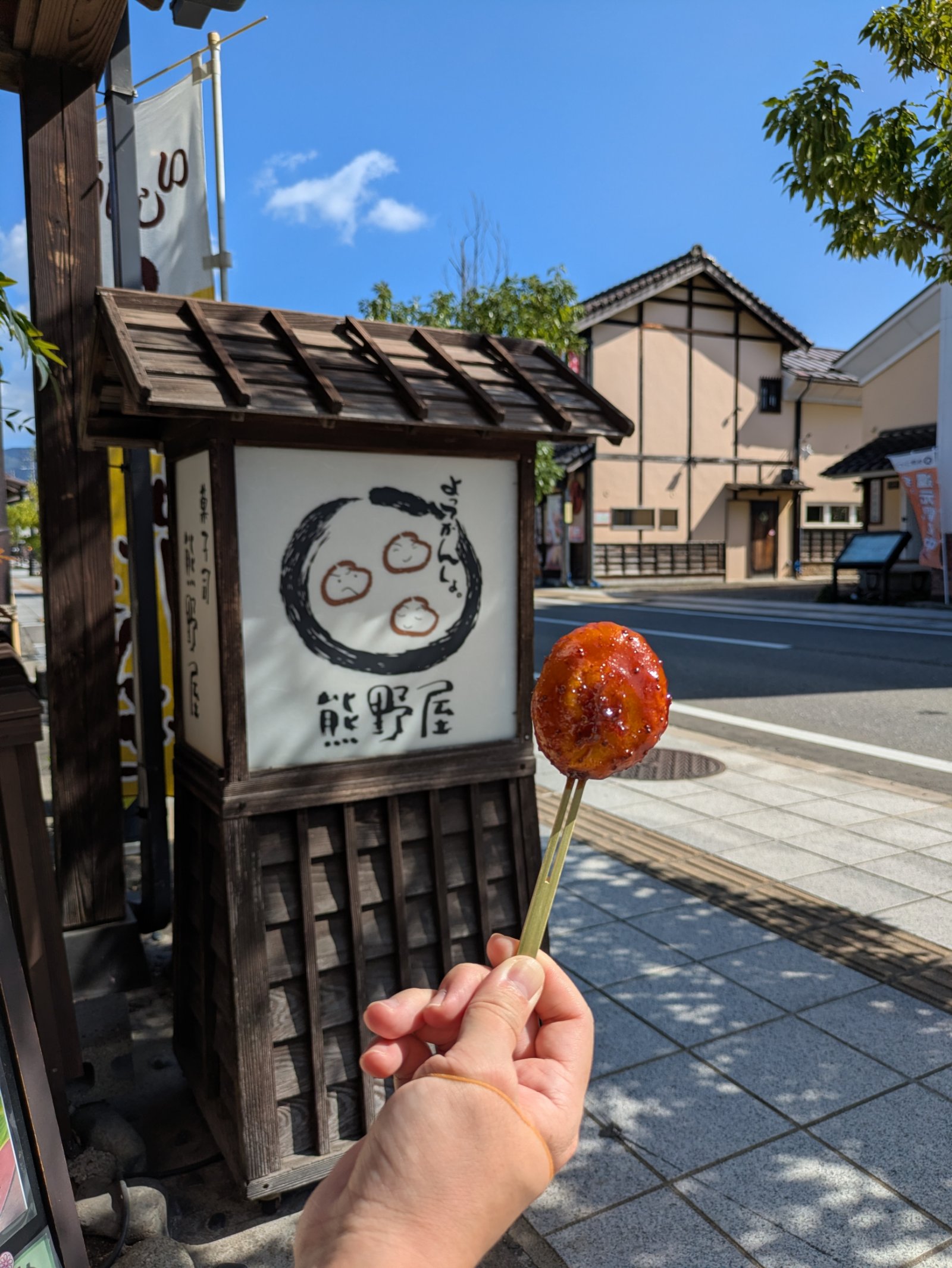 熊野屋の看板の前で手に持った串刺しのしんごろう餅、晴れた七日町の通り