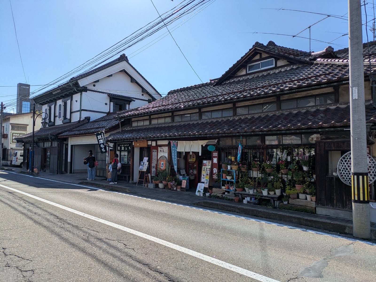 瓦屋根の古い町家と白壁の蔵造りの建物が並ぶ晴れた日の七日町通り。軒先に鉢植えが並び、「豆腐もち」の旗の前に人が立つ