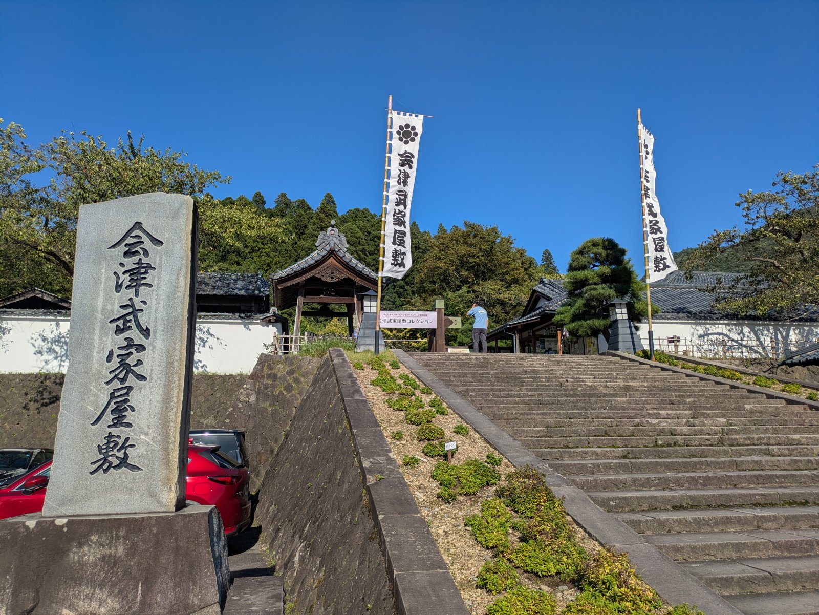 「会津武家屋敷」と刻まれた石碑と、のぼり旗が立つ入口へ続く石段。晴天の青空の下