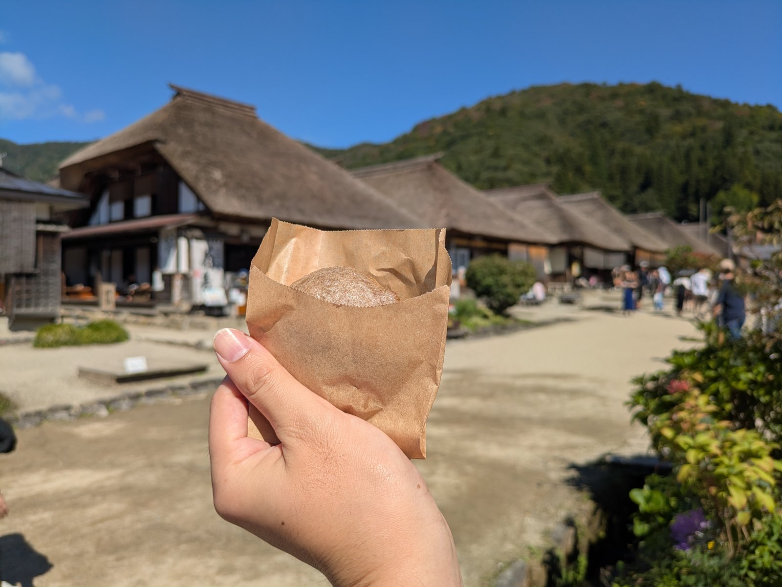 大内宿の茅葺屋根の家並みを背に、紙袋に入ったそば粉のまんじゅうを手に持つ様子