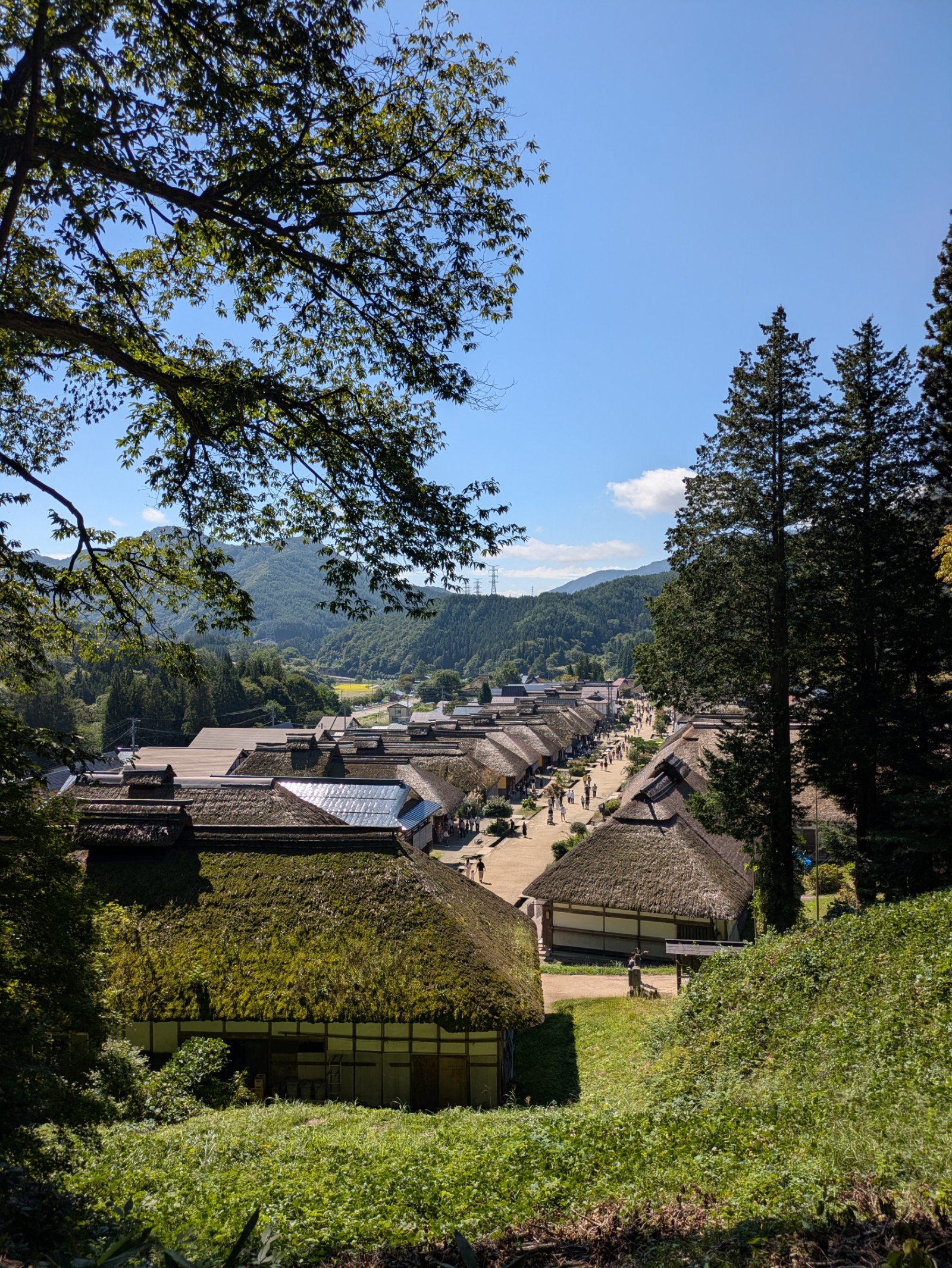 高台から見下ろす大内宿の茅葺き屋根の家並みと、夏の青空の下に続く山々