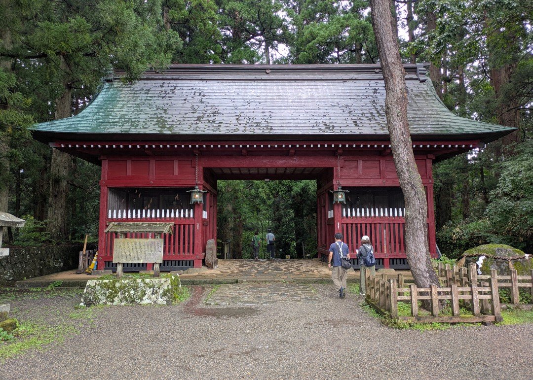 杉木立に囲まれた朱塗りの門と、門をくぐって参道へ進む参拝者たち