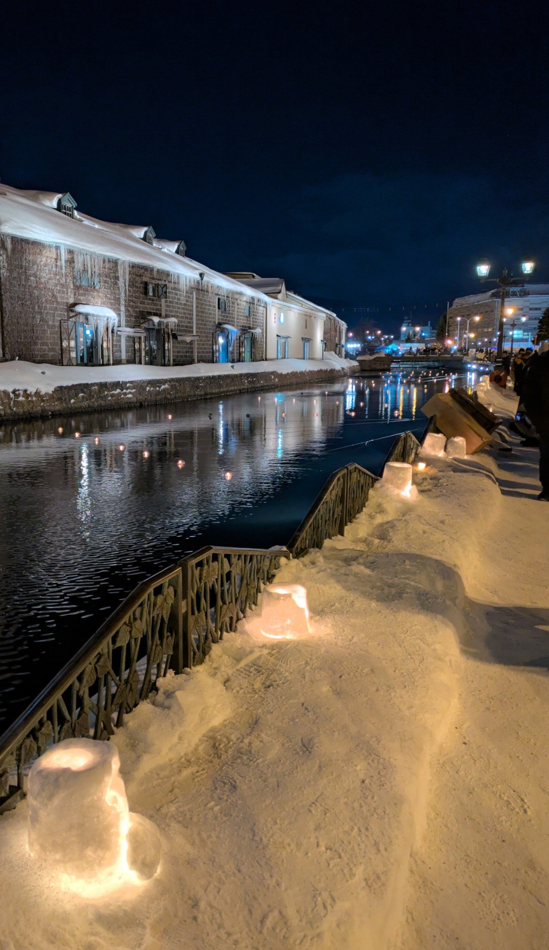 夜の小樽運河沿いの雪道に並ぶ灯籠と、水面に浮かぶ灯り、雪をかぶった石造倉庫