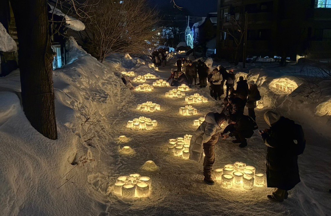 雪の小道に並ぶ灯りのともったスノーキャンドルと、写真を撮る見物客(おたる雪あかりの路・旧手宮線会場)