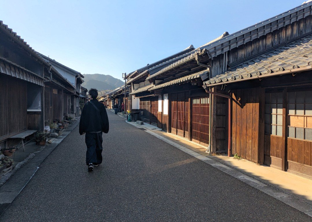 晴れた日の関宿、木造の町家が両側に並ぶ一本道を歩く黒い服の人
