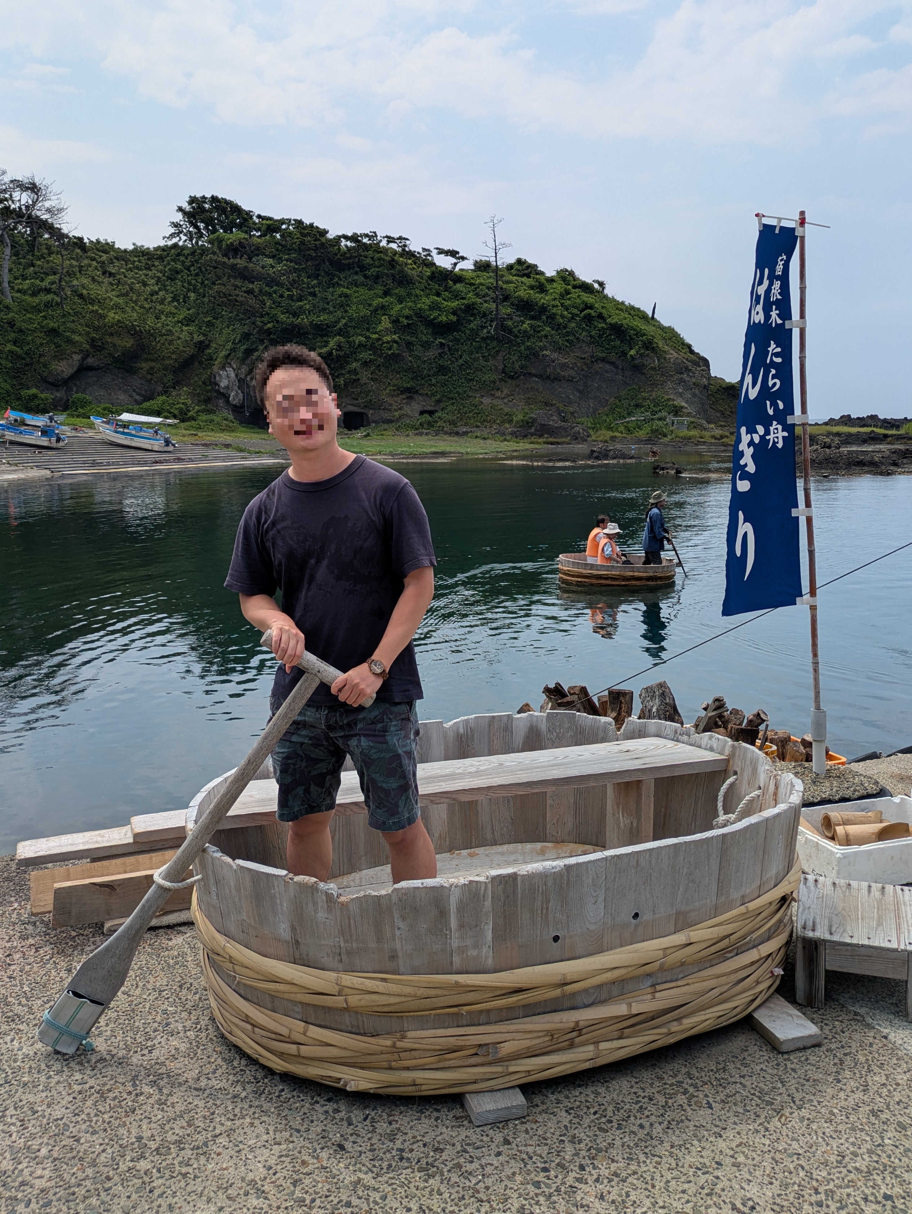 宿根木の岸辺でたらい船に立ち櫂を握る男性、奥の入り江にもう一艘のたらい船
