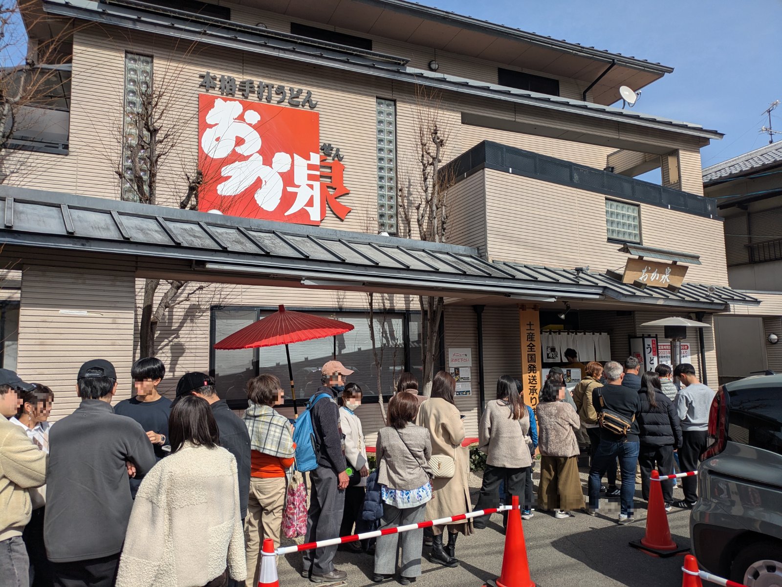 「本格手打うどん おか泉」の看板を掲げた店の前に並ぶ大勢の客の行列、晴れた日