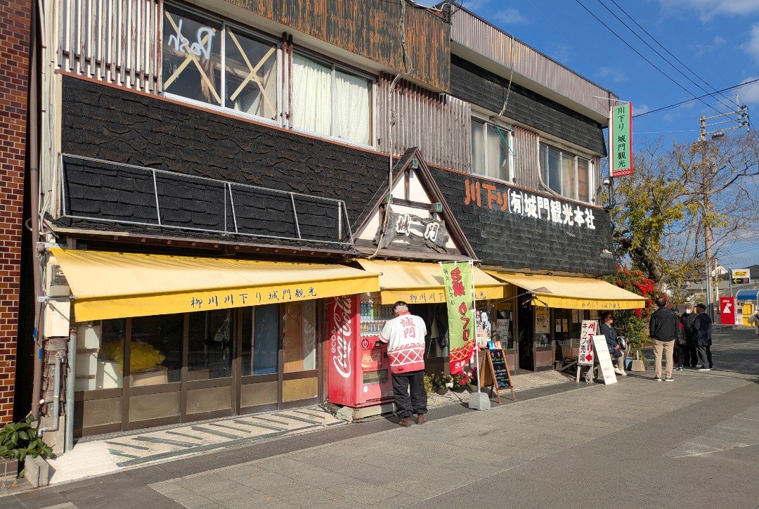 黄色い日よけが並ぶ柳川川下り城門観光の店舗と、法被姿の男性