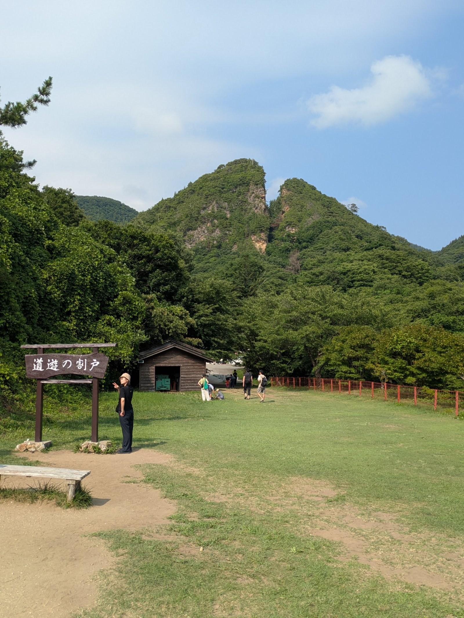 「道遊の割戸」の看板の先にそびえる、山頂が二つに割れた緑の山と夏の芝生広場